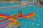 ein junges Mädchen paddelt grinsend auf mehreren, bunten Schwimmstäben durchs Becken