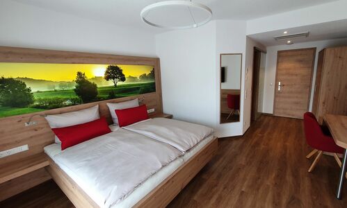 Der Adler - Hotelzimmer in hellem modernen Holz. Im Kopfteil des Bettes ist ein Leuchtbild. Es schweift der Blick über die Felder mit aufsteigendem Nebel bei Sonnenaufgang.